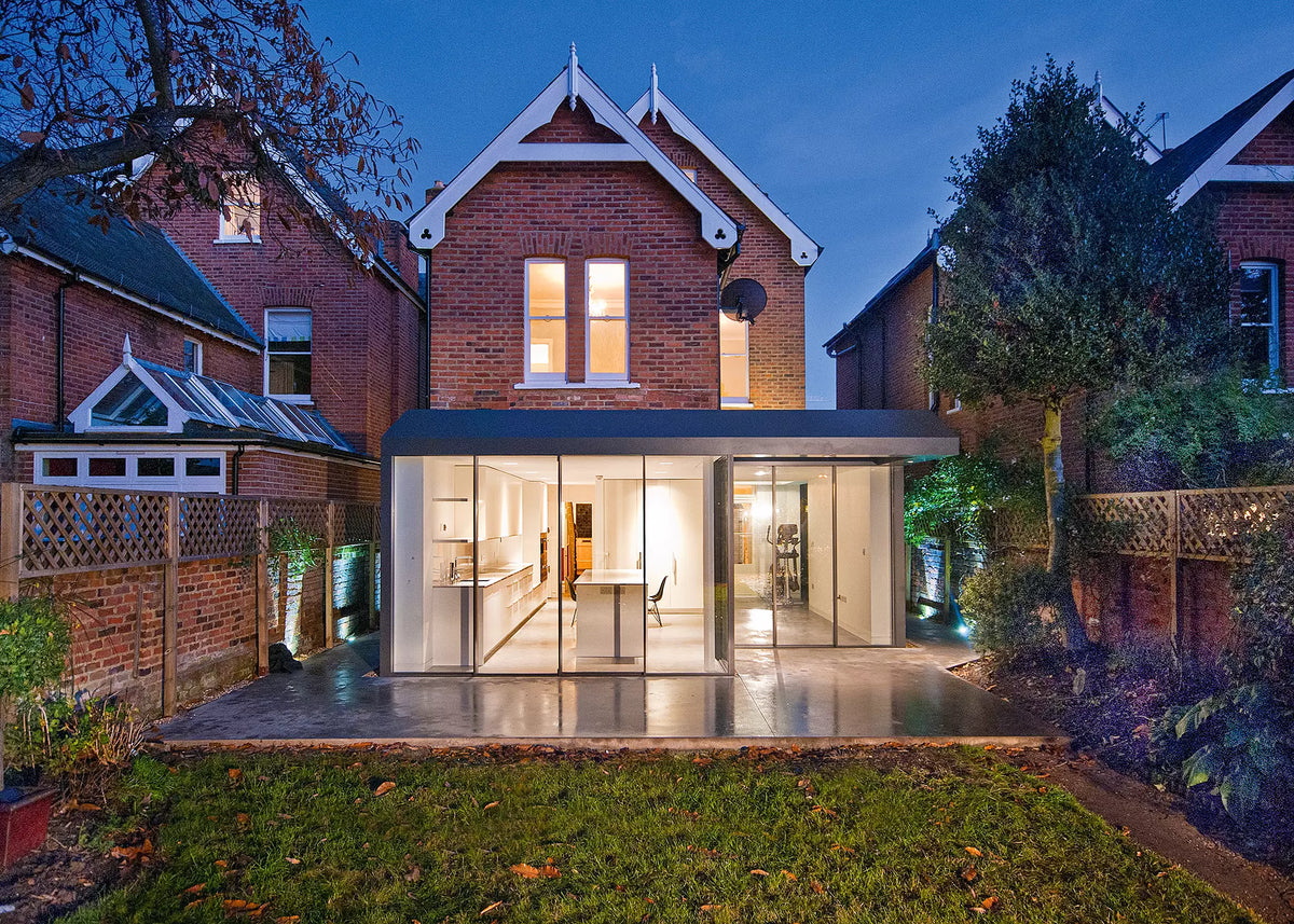 A modern glass extension with a dark roof over a brick house at dusk, showing a kitchen and dining area illuminated from within, with a lawn and neighboring houses in the background.